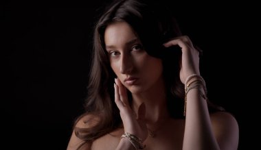 Uzun kahverengi saçlı, bilezik ve kolye takan, saçına ve yüzüne dokunan genç bir kadının stüdyo portresi.