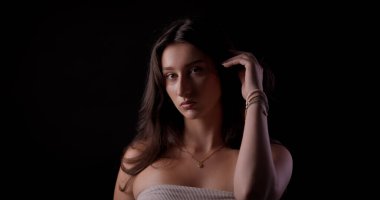 Uzun kahverengi saçlı, kolye ve bilezik takan, siyah arka planda saçlarına dokunan genç bir kadının stüdyo portresi.