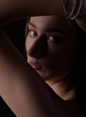 Kollarını kaldırmış, bilezik takmış, dramatik ışıklandırmayla aydınlatılmış, gizemli ve cezbedici bir şey yaratan genç bir kadının stüdyo resmi.