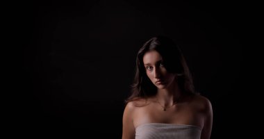 Uzun kahverengi saçlı, beyaz bluz ve kolye takan, siyah arkaplanlı genç bir kadının stüdyo portresi.