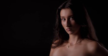 Uzun kahverengi saçlı, siyah arka planda duran altın kolye takan çekici bir genç kadının stüdyo portresi.