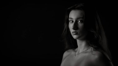 Uzun saçlı, kolye takan, siyah beyaz bir kadın portresi. Stüdyoda siyah arka planda poz veriyor.