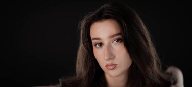 Stüdyo portresi. Uzun kahverengi saçlı güzel bir kadın. Koyu bir arka planda poz veriyor.