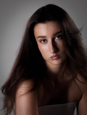 Uzun kahverengi saçlı, çıplak omuzlu, beyaz bluzlu, ciddi bir genç kadının stüdyo portresi.