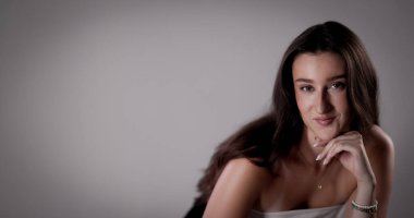 Uzun kahverengi saçlı, elleri çenesinde gri arka planda poz veren beyaz bir kadının stüdyo portresi.