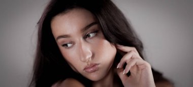 Uzun kahverengi saçlı güzel bir kadının portresi. Yüzüne eliyle dokunuyor ve başka tarafa bakıyor.