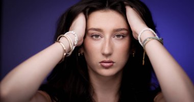 Uzun kahverengi saçlarını tutan, mor arka planda bilezik takan genç bir kadının stüdyo portresi.