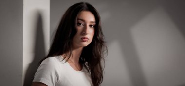 Uzun kahverengi saçlı, beyaz tişörtlü ve stüdyo ortamında beyaz bir duvara yaslanmış ciddi bir genç kadının portresi.