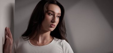 Uzun kahverengi saçlı, beyaz tişörtlü, beyaz duvara yaslanmış üzgün genç bir kadın başka tarafa bakıyor.