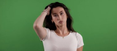 Sıkılmış bir ifadeyle genç bir kadın yeşil arka planda saçlarına dokunuyor.