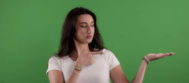 Yeşil ekran arka planında şüpheli bir yüz ifadesiyle açık elini gösteren genç bir kadın.