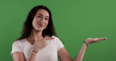 Genç bir kadın açık elini göstererek yeşil ekranda bir şey gösteriyor.