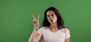 Stüdyo portresi genç bir kadının komik bir yüz ifadesi ve yeşil perde arkasında zafer işaretini gösteriyor.