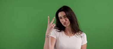 Genç kadın parmaklarıyla zafer işareti gösteriyor ve yeşil arka planda komik bir ifade takınıyor.