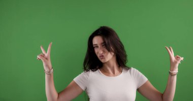 İki eliyle barış işareti yapan ve yeşil ekran arka planında somurtan genç bir kadın.