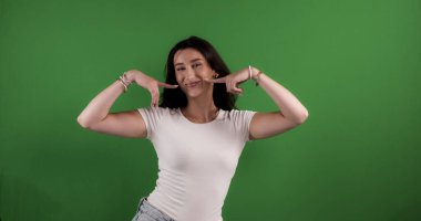 Genç bir kadın işaret parmaklarıyla yanaklarını gösteriyor, krom anahtar arka planda gülümsüyor.