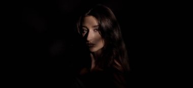 Gölgelerden çıkan uzun kahverengi saçlı büyüleyici genç bir kadının stüdyo portresi ilgi çekici ve gizemli bir atmosfer yaratıyor.