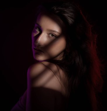 Uzun kahverengi saçlı güzel bir kadının moda portresi. Mor bir ışıkla aydınlatılmış. Yüzünde ve omzunda çizgili gölgeler yaratıyor.
