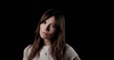 Uzun kahverengi saçlı, beyaz tişörtlü, koyu bir arka plana karşı ciddiyetini ifade eden genç bir kadının stüdyo portresi.