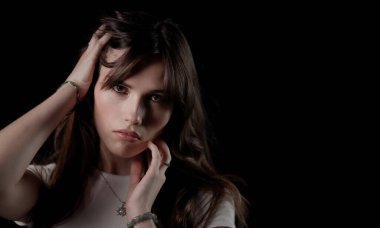 Uzun kahverengi saçlı ve takılı genç bir kadının stüdyo portresi, koyu bir arka planda saçına ve yüzüne dokunuyor.