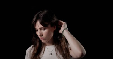 Saçına dokunan, siyah arka planda bilezik ve kolye takan genç bir kadının stüdyo portresi.