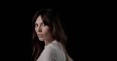 Omuzlarının üzerinden siyah bir arka plana bakan uzun kahverengi saçlı güzel bir genç kadının portresi.