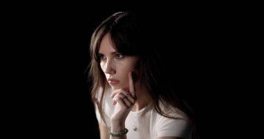 Elleri çenesinde düşünen, siyah arka plana bakan genç bir kadın.