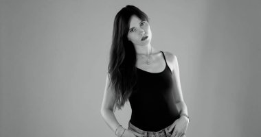 Uzun kahverengi saçlı, siyah bluzlu ve kotlu elleri cebinde poz veren genç bir kadının stüdyo portresi.