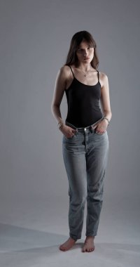 Stüdyoda çıplak ayakla poz veren ve elleri cebinde kot pantolon ve kolsuz bluz giyen genç bir kadının portresi