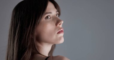 Uzun kahverengi saçlı, gri arka planda düşünceli bir ifadeyle yukarı bakan genç bir kadının profili.