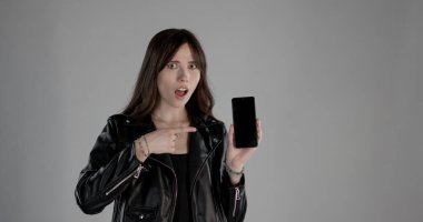 Akıllı telefonu boş ekranla gösteren ve şaşırmış bir ifadeyle işaret eden genç bir kadın.