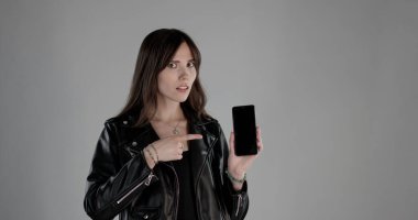 Genç bir kadın gri arka planda boş bir ekranla akıllı telefonu işaret ediyor.