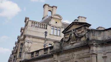 Cambridge 'deki tarihi bir binanın düşük açılı görüntüsü, detaylı taş oymalar, bacalar ve mimari özellikler sergileniyor.