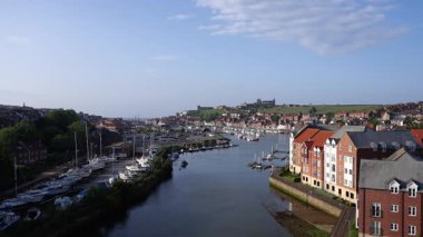 Kuzey Yorkshire, İngiltere 'deki Whitby limanının manzarasında çok sayıda tekne, ikonik Whitby Manastırı ve açık mavi gökyüzü altındaki şehir manzarası var.
