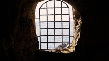 Karanlık bir mağaranın içindeki parmaklıklı bir pencereden deniz manzarası hapsedilme, kaçış ve özgürlük özlemi kavramlarını çağrıştırıyor.