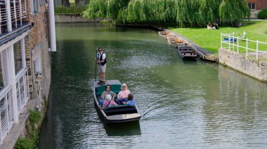 CAMBRIDGE, İngiltere - 11 Haziran 2025 - Gondolier Cambridge, İngiltere 'deki Cam nehri boyunca turistleri tekmeliyor, söğütler ve demirlemiş teknelerin yanından geçiyor