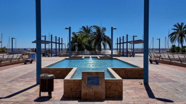 Marbella, İspanya marinasındaki bir çeşmenin manzarası, palmiye ağaçları, mavi direkler ve açık mavi gökyüzü - MARBELLA, SPAIN - 30 Mart 2025