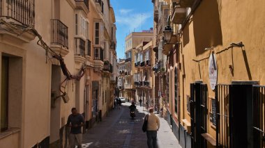 İnsanlar Cadiz, İspanya 'da güneşli bir günde, 26 Mart 2025' te, Cadiz, İspanya 'da, dar bir sokakta yürüyorlar ve scooter sürüyorlar.