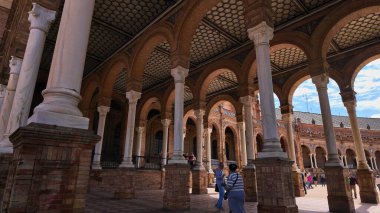 Plaza de Espana, Seville, İspanya 'da beyaz mermer sütunların ve tuğla kemerlerin altında yürüyen turistler - SEVILLE, İspanya - 25 Mart 2025