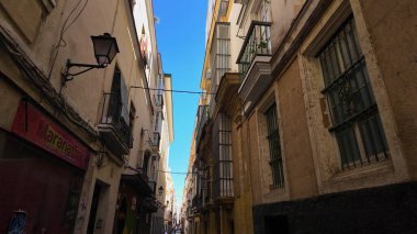Cadiz 'in eski kasabasında dar bir caddeyi karakterize eden geleneksel mimari, açık mavi gökyüzü altında balkonlar, parmaklıklı pencereler ve sokak lambaları - CADIZ, SPAIN - 26 Mart 2025