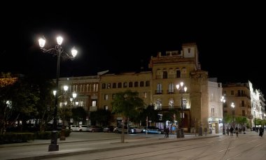 Sevilla 'da gece sokak lambalarıyla aydınlatılmış, park halindeki araçlar ve yürüyen insanlar - SEVILLE, SPAIN - 25 Mart 2025