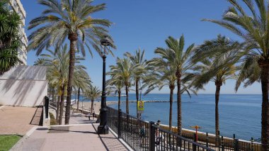 Güneşli bir yaz gününde İspanya 'nın Marbella kentindeki Akdeniz manzaralı palmiye ağaçlarıyla kaplı gezinti güvertesinde yürüyen turistler - MARBELLA, İspanya - 30 Mart 2025