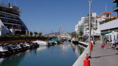 Limanda güneşli bir günün tadını çıkaran insanlar, limanda demirlemiş tekneler, restoranlar ve yaya köprüsü ile suya yansıyan GIBRALTAR, İngiltere - 31 Mart 2025