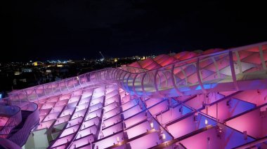 Metropol Şemsiyesi 'nin yükseltilmiş geçidi gece mor ve mavi ışıklarla aydınlatılmış ve arka planda Seville şehri manzarası - SEVILLE, SPAIN - 25 Mart 2025