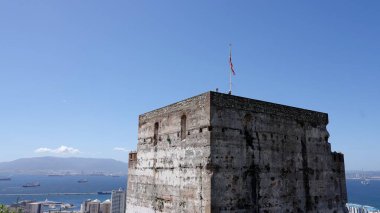 Mağribi Kalesi rüzgarda dalgalanan Cebelitarık bayrağını sergiliyor, mavi gökyüzü ve deniz arka planda