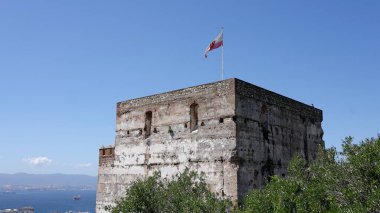 Mağribi Kalesi rüzgarda dalgalanan Cebelitarık bayrağını sergiliyor, mavi gökyüzü ve deniz arka planda