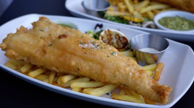 Lezzetli haşlanmış balık ve patates kızartması, limon dilimleri, tartar sosu ve beyaz tabakta yeşil sos.