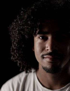 Karanlıktan çıkan kıvırcık saçlı ve keçi sakallı Afro-Amerikan bir adamın portresi.