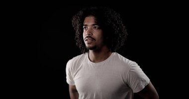 Siyah bir arka plana bakan kıvırcık saçlı ve beyaz tişörtlü Afro-Amerikan bir adamın portresi.