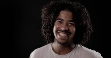 Siyah arka planda gülümseyen kıvırcık saçlı, neşeli, Afro-Amerikan bir adamın portresi.